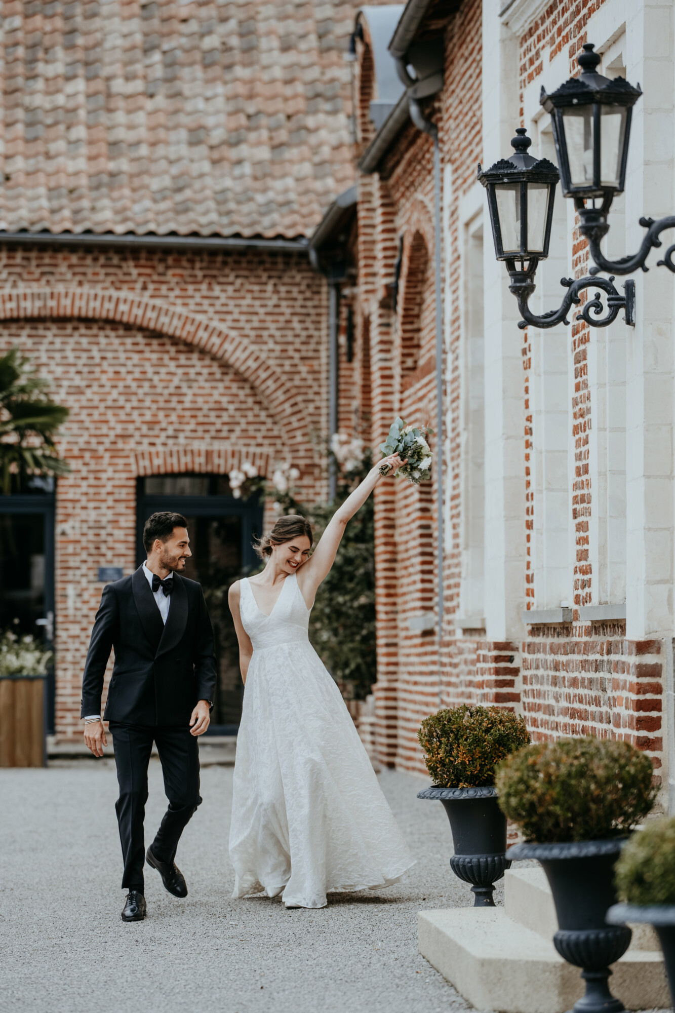Couple de mariés marchant dans la cour d'honneur du Domaine de la Traxène entre les façades en briques.