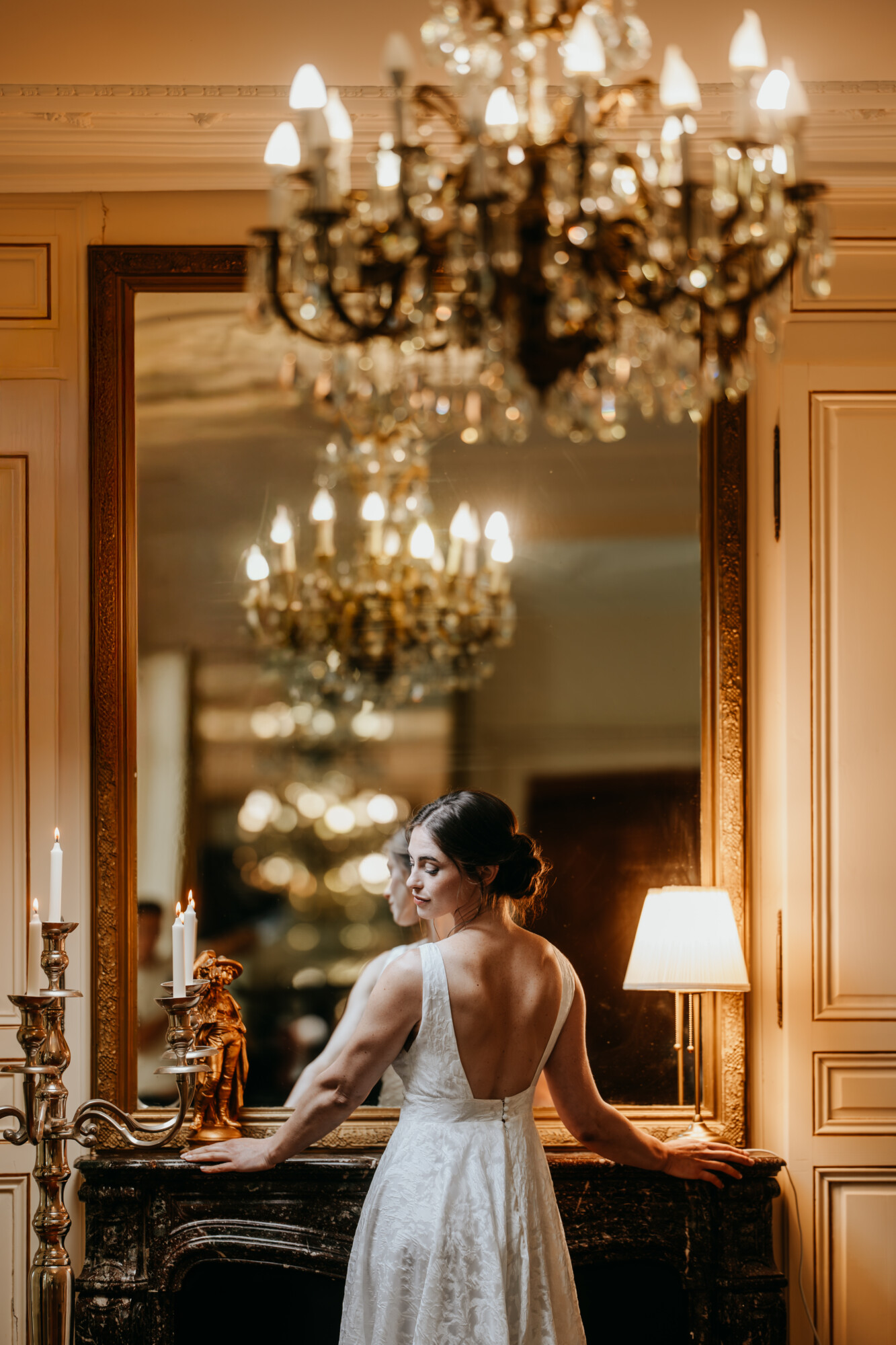 Copie de Julien Briche Photographe-6 – Copie Mariée de dos devant une cheminée en marbre, un grand miroir ancien et un lustre en cristal dans un salon de caractère au Domaine de la Traxène.