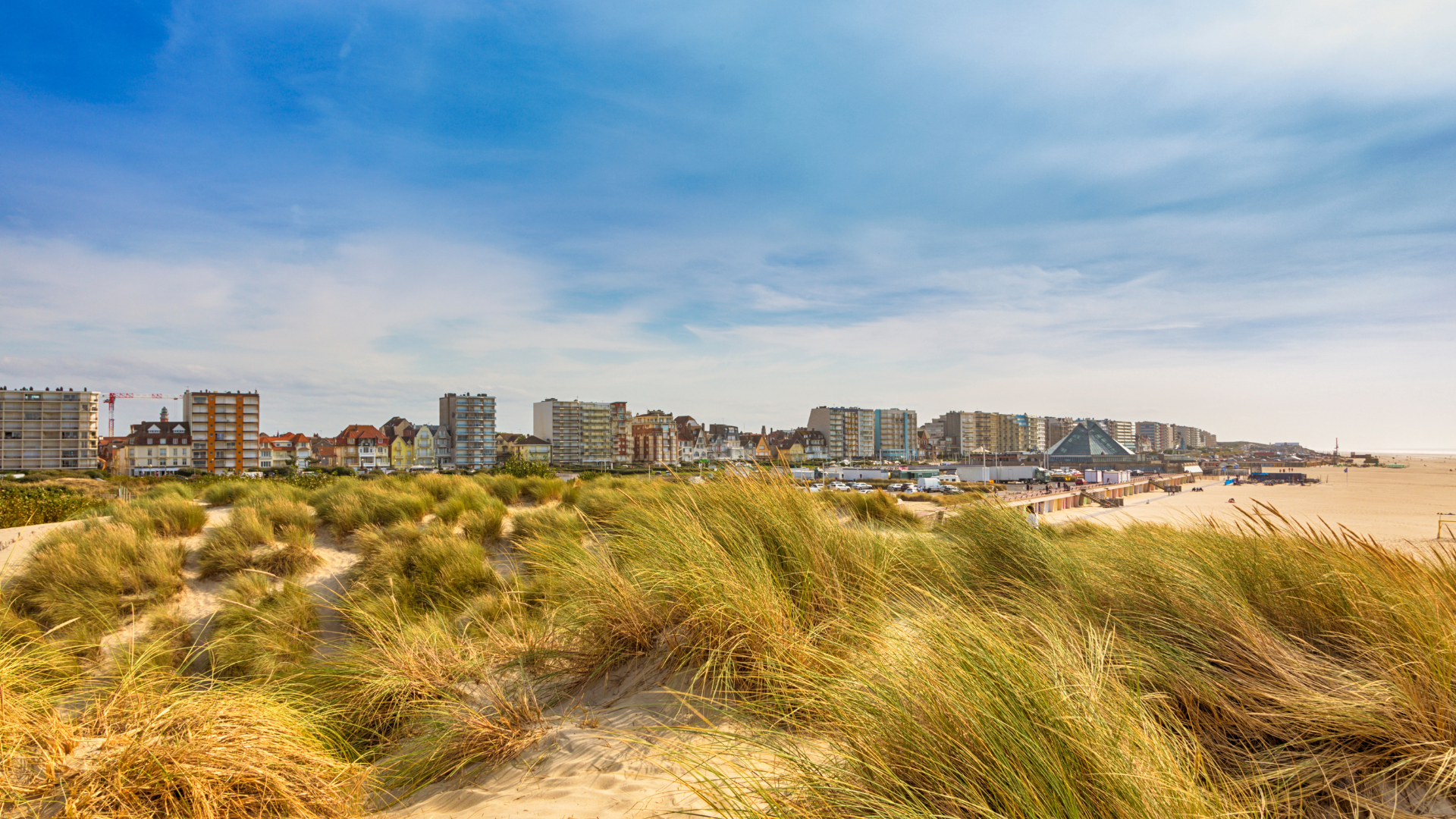 Vue panoramique du Touquet-Paris-Plage depuis les dunes, avec la plage de sable, les oyats et le front de mer sous un ciel bleu.